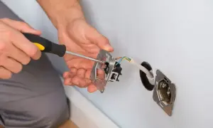 Ręce majsterkowicza przykręcają śrubokrętem mechanizm gniazdka. Widać przewody elektryczne i puszkę instalacyjną.