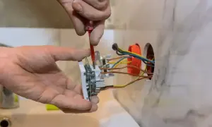 Ręce majsterkowicza dokonują podłączenia gniazdka. Widać przewody elektryczne wychodzące z puszki instalacyjnej.