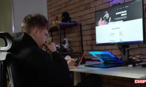 Młody mężczyzna w okularach sprawdza zasięg sieci mobile vikings na telefonie, siedząc przy biurku z otwartym laptopem i monitorem.