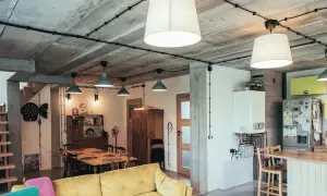 Nowoczesne wnętrze z widoczną instalacją elektryczną natynkową na betonowym suficie. Wygodna kanapa, stół jadalniany i kuchnia.