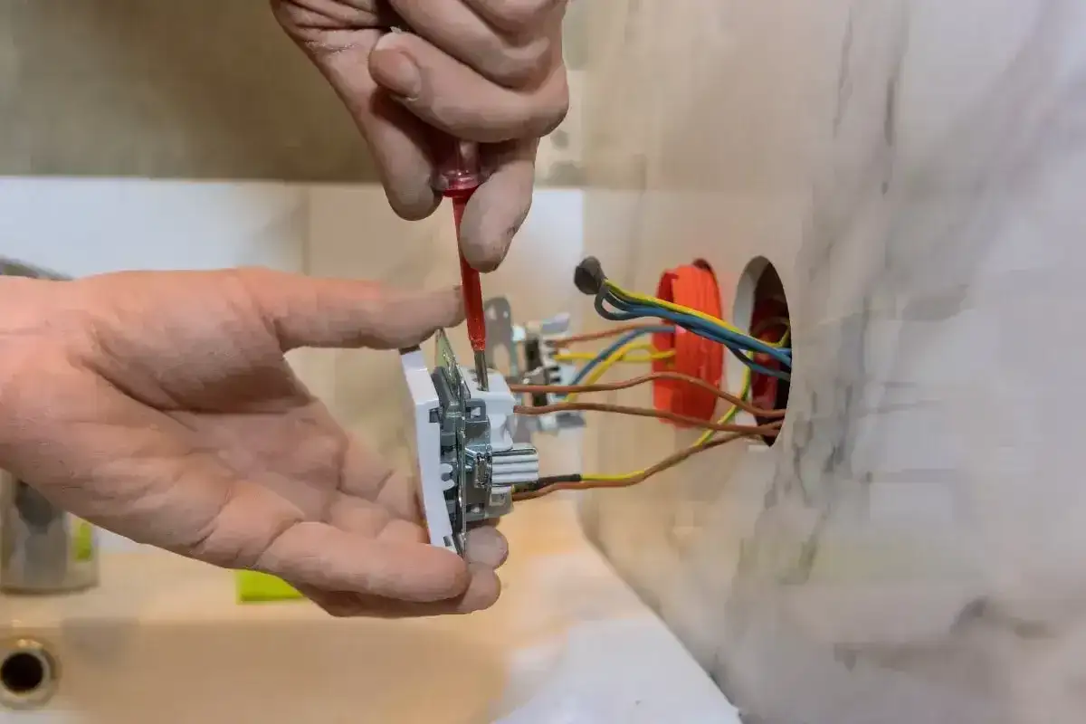 Ręce majsterkowicza dokonują podłączenia gniazdka. Widać przewody elektryczne wychodzące z puszki instalacyjnej.