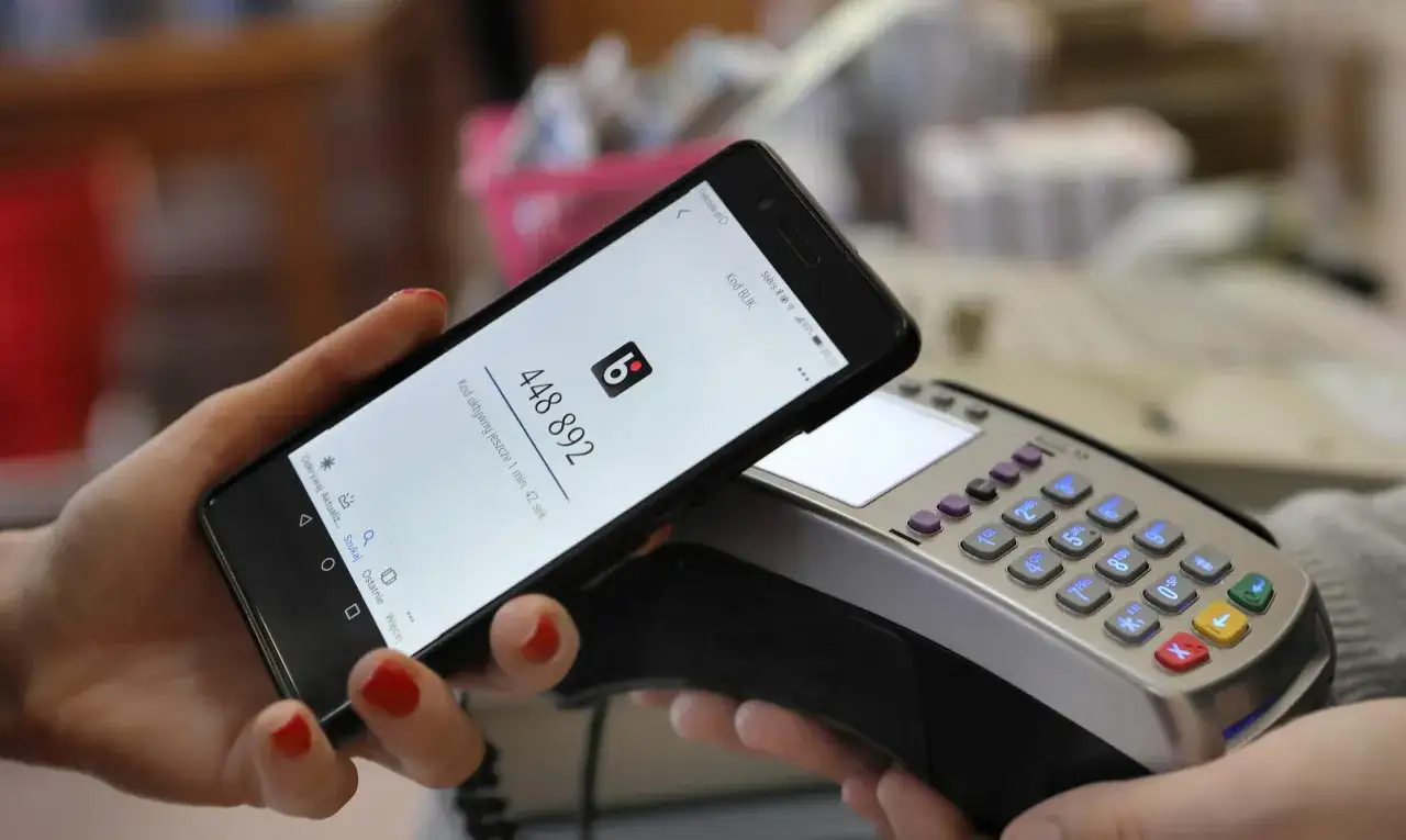 Kobieta zbliża telefon do terminala płatniczego, pokazując kod BLIK. Tak łatwo można płacić telefonem.