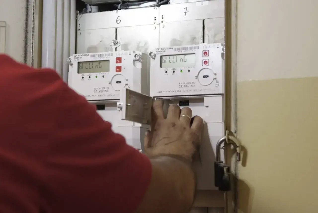 Ręka w czerwonej bluzce otwiera klapkę licznika energii elektrycznej, na którym widać cyfry. To część systemu, który może wykorzystywać kody OBIS.